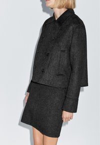 Kvinde iført en mørkegrå uld, dobbeltradet jakke og matchende nederdel, står foran en ensfarvet lys baggrund.