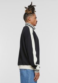 Sort sweatshirt med hvide striber, høj krave og ribbede manchetter. Har en afslappet pasform og blød tekstur, parret med lyseblå denim.