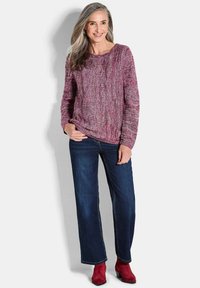 Burgunderfarbener Strickpullover mit strukturiertem Zopfmuster. Kombiniert mit blauen Jeans im geraden Schnitt und roten Stiefeletten.
