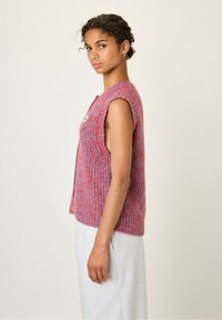 Žena s kučeravými vlasmi v textúrovanej bezrúškovej červenej a fialovej pletenej vesty, v kombinácii so svetlo šedými nohavicami, stojí na pozadí bez vzoru.