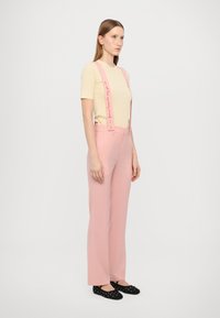 Pink trousers with wide legs and adjustable suspenders featuring decorative text; paired with a fitted beige t-shirt and black dotted flats.