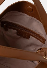 Interior de un bolso de cuero marrón texturizado con forro beige, un bolsillo con cremallera y un parche de logo con la inscripción "LIU JO".