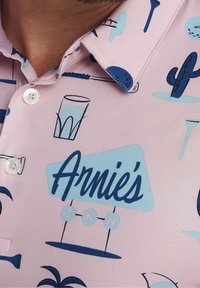 Polo rose clair avec un motif de boissons tropicales bleues, de cactus et le texte « Arnie's INN ». Comprend des boutons blancs et un col.