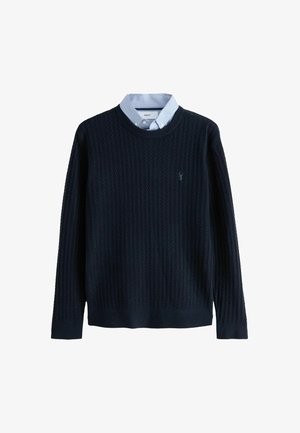 Pull en maille côtelée bleu marine avec un col rond et des manches longues, superposé à une chemise à col bleu clair. Logo discret sur la poitrine.