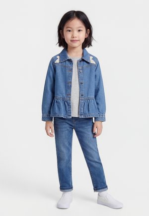 Νεαρή κοπέλα στέκεται φορώντας μπλε τζιν μπουφάν με σχέδια αρκούδας, γκρι μπλούζα, μπλε τζιν παντελόνι, λευκές κάλτσες και λευκά πάνινα παπούτσια σε λευκό φόντο.