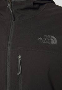Czarna kurtka softshell z przodu na zamek, bocznymi kieszeniami i logo na klatce piersiowej. Materiał wydaje się teksturowany i trwały.