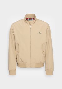 Beige bomber bunda se zipem v přední části, žebrovanými manžetami a kostkovanou podšívkou. Obsahuje boční kapsy a malou zelenou nášivku na hrudi.