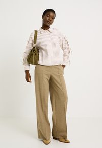 Femme portant un pantalon large beige, une chemise rayée blanche et beige avec des liens aux manches, des bottes beiges et portant un sac à bandoulière vert olive.