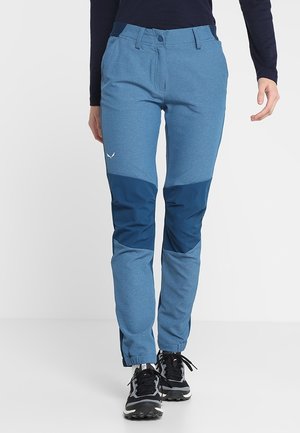 Personne portant un pantalon d'extérieur bicolore bleu avec des poignets élastiques et des chaussures de sport noires, debout devant un fond blanc uni.