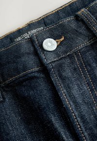 Tmavé džínsy z denimu s medeným prešívaním, so strieborným kovovým gombíkom a viditeľnou značkovou etikettou na páse.