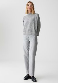 Grijs, textuurtrui met lange mouwen, gecombineerd met lichtgrijze rechte jeans en zwarte instappers. Effen achtergrond.
