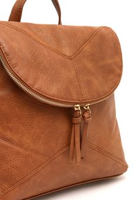 Sac à dos en cuir marron avec un rabat arrondi, une fermeture éclair dorée et deux glands en cuir. Présente une surface texturée et des détails de couture géométriques.