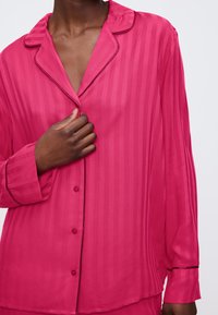Rosa randig pyjamasöverdel med notched krage, långa ärmar, knappar framtill och en slät yta; har kontrasterande pipingdetaljer.