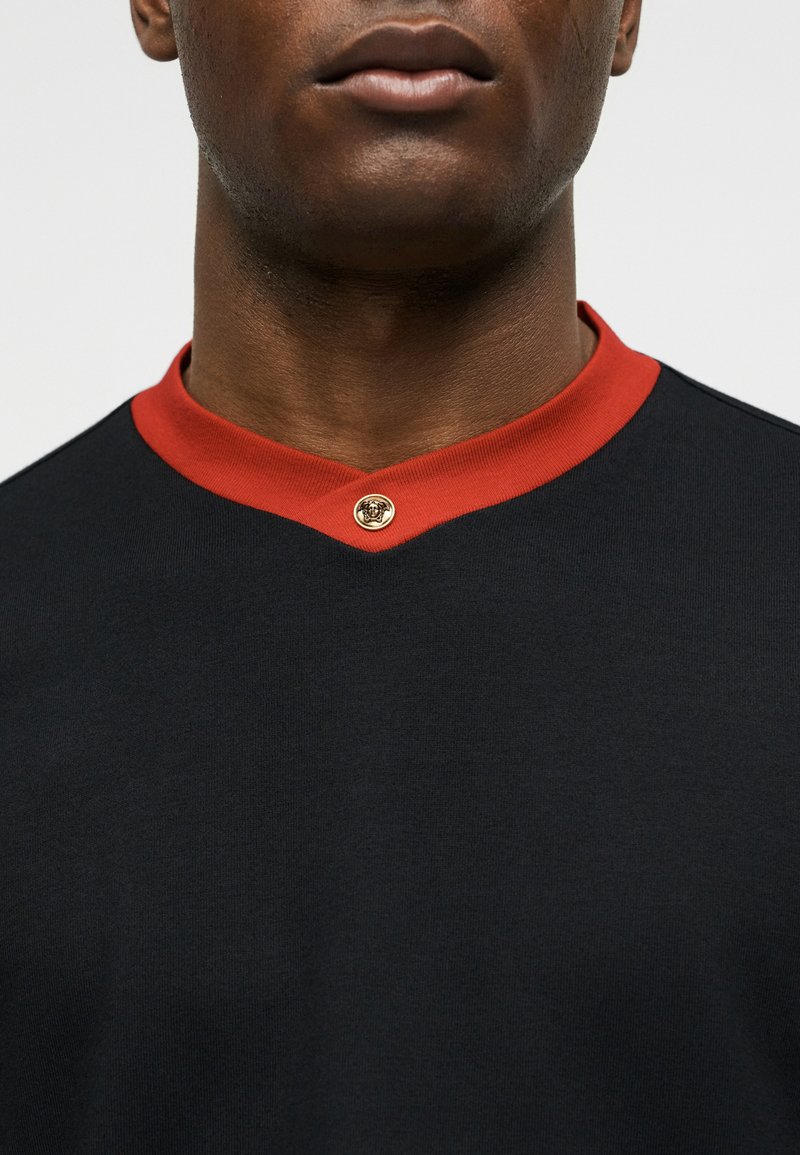 Gros plan d'une personne portant une chemise noire avec un col rouge et un petit bouton doré au centre devant.