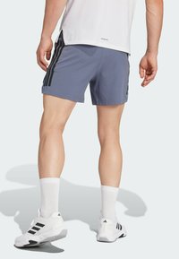 Pantaloni sportivi da uomo in azzurro chiaro con strisce laterali nere. Indossati con calze bianche e sneakers bianche con dettagli neri, che mettono in mostra gambe muscolose.