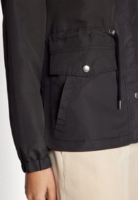 Giubbotto leggero nero con chiusura a zip frontale, polsini elasticizzati e due tasche frontali con chiusura a pressione. Tessuto liscio, design casual.