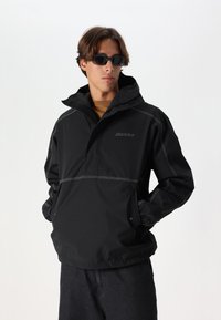 Black waterproof jacket with a hood, featuring a half-zip front, elastic cuffs, contrast stitching, and side pockets. Brand logo displayed.