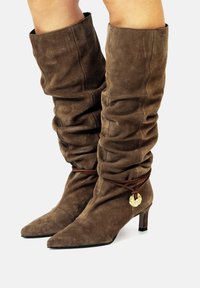 Botas de ante marrón hasta la rodilla con un diseño desaliñado, puntera puntiaguda y un acento metálico asegurado por un cordón marrón. Tacón bajo tipo bloque.