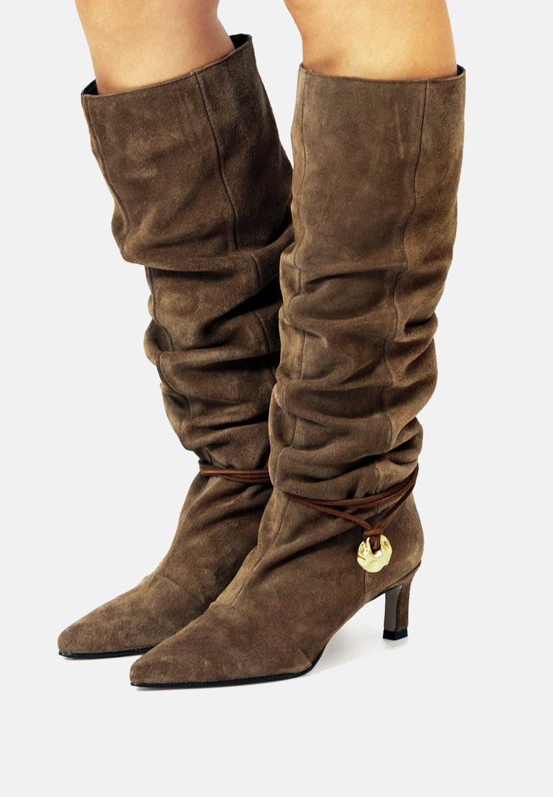 Botas de ante marrón hasta la rodilla con un diseño desaliñado, puntera puntiaguda y un acento metálico asegurado por un cordón marrón. Tacón bajo tipo bloque.