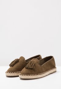 Braune Wildleder-Pantoletten mit einem Quastenakzent, gewebtem Jute-Mittelsohle und glatter Textur. Abgerundete Zehenform und flexible Sohle.