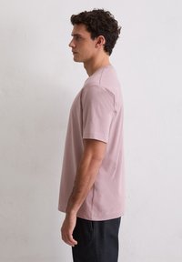 Młody mężczyzna stojący w profilu, ubrany w gładką, jasnoróżową koszulkę z krótkim rękawem oraz ciemne spodnie, na tle białej, teksturowanej ściany.