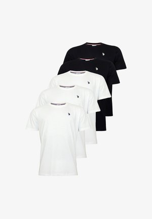Jeune homme debout, portant un t-shirt blanc à manches courtes avec un petit logo, un jean bleu et des baskets noires avec des lacets blancs sur un fond clair.
