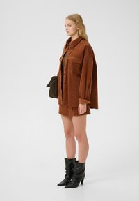 Veste chemise en corduroy marron avec deux poches avant, assortie à une mini-jupe marron. Des bottes noires à talons complètent la tenue. Tissu texturé.
