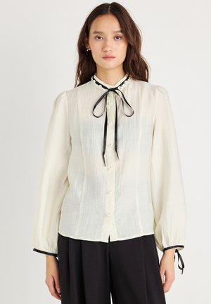 Krém színű blúz hosszú puffos ujjal, fekete szalaggal a nyakrészen, gyöngygombokkal és fekete díszítéssel az ujjak végén. Puha, textúrált anyag.