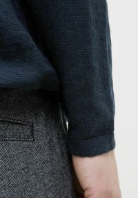 Ciemnoniebieski sweter dziany o teksturowanym wykończeniu, ukazujący zbliżenie rękawa oraz wzorzystą szaro-skalną tkaninę spodni w jodełkę.