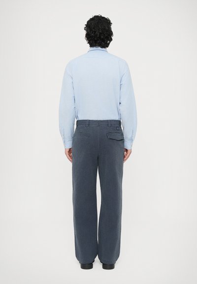 Μπλε πουκάμισο με μακριά μανίκια και γιακά με κουμπιά, συνδυασμένο με φαρδιά, σκούρα γκρι υφασμάτινα παντελόνια. Ορατές πίσω τσέπες και ελαφριά γυαλάδα στο ύφασμα.