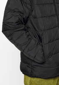 Black puffer jacket with quilted sections, zippered pockets, and a smooth finish, worn over olive green pants.