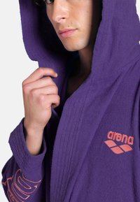 Lilla hættekjole lavet af blødt stof, med en tekstureret overflade, og med et orange "arena" logo på brystet og ærmet.