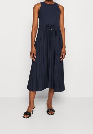Persoon draagt een mouwloze marineblauwe midi-jurk met geplooide rok en koord in de taille, gecombineerd met zwarte sandalen met hoge hakken.