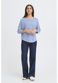 Gaiši zils tekstūrēts adījuma tops ar 3/4 piedurknēm, apvienots ar tumši zilām platām biksēm un baltām čēpēm, stāv pret vienkāršu fonu.