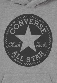 Šedá mikina s kapucí s kruhovým černým logem se hvězdou. Text zahrnuje "Converse," "Chuck Taylor," a "All Star" v různých písmových stylech.