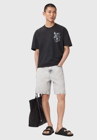 Άνδρας με μαύρο T-shirt με σχέδιο, ανοιχτόχρωμα τζιν σορτς, μαύρα σανδάλια, κρατώντας μεγάλη μαύρη τσάντα, στέκεται μπροστά σε απλό φόντο.