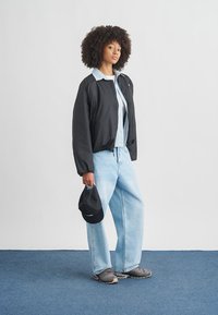 Svart bomberjakke med lyseblå krage, kombinert med lyseblå vide jeans og grå joggesko. Holder en svart caps.