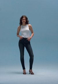 Fehér ujjatlan felső, sötét skinny farmernadrág és fekete magassarkú szandál, világoskék háttér előtt állva. Sima anyag és testhezálló fazon.