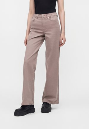 Personne portant un pantalon large marron clair, un haut noir et des chaussures compensées noires épaisses, debout sur un fond uni.
