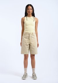 Top de tirantes amarillo claro con textura en relieve, pantalones cortos de mezclilla beige con bolsillos frontales y un ligero dobladillo, combinados con zapatillas grises y calcetines de tobillo estampados.