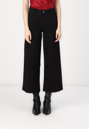 Vrouw draagt zwarte, wijd uitlopende, cropped broek, zwarte laarzen met spitse neus en een rood topje, staand tegen een witte achtergrond.