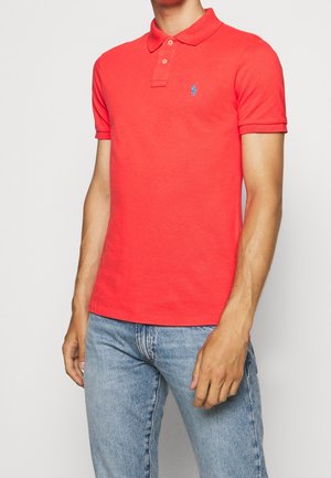 Mann trägt ein leuchtend rotes Polohemd mit kurzen Ärmeln und blauem Logo auf der Brust sowie hellblaue Jeans, steht vor einem schlichten Hintergrund.