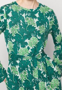 Abito a maniche lunghe con scollo rotondo, caratterizzato da un motivo floreale verde su uno sfondo azzurro chiaro, vita cintrata e tessuto strutturato.