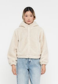 Nő, aki krémszínű fleece zipzáras kapucnis pulóvert visel világoskék farmernadrággal, egy sima fehér háttér előtt áll.