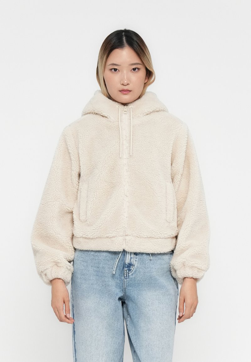Vrouw in een crèmekleurige fleece hoodie met rits en capuchon, met lichtblauwe denim jeans, staande tegen een effen witte achtergrond.