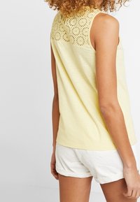 Geel mouwloos topje van zacht materiaal, met een gedetailleerd bloemenoogletontwerp op de rug. Gecombineerd met witte shorts.
