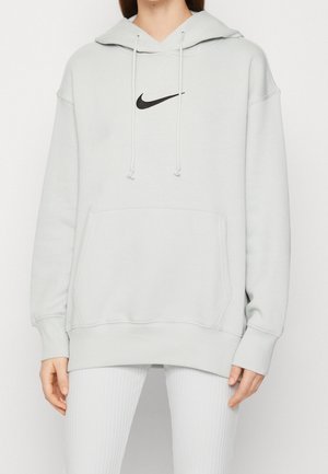 Kvinde iført lys grå oversized Nike-hoodie med sort swoosh-logo og hvide ribstrikkede leggings, stående mod en ensfarvet baggrund.