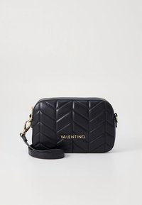 Svart quiltad läderclutch från Valentino med guldfärgad logotyp och avtagbar handledsrem på ljus bakgrund.