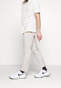 Krämfärgad kortärmad skjorta, ljusbeige randiga byxor och vita Nike-sneakers med svarta detaljer, stående mot en neutral bakgrund.