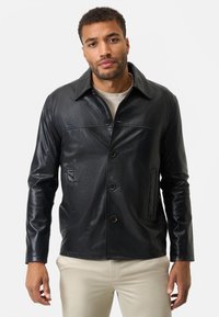 Schwarze Lederjacke mit einem Kragen, vorderem Knopfverschluss, zwei Seitentaschen und glatter Textur. Über einem hellen Pullover getragen.